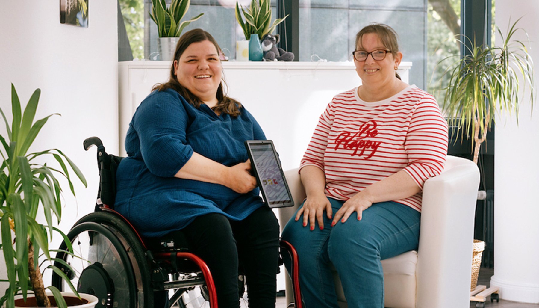 Eine Rollstuhlfahrerin mit Tablet und eine Frau sitzen sich gegenüber, beide lachen in die Kamera