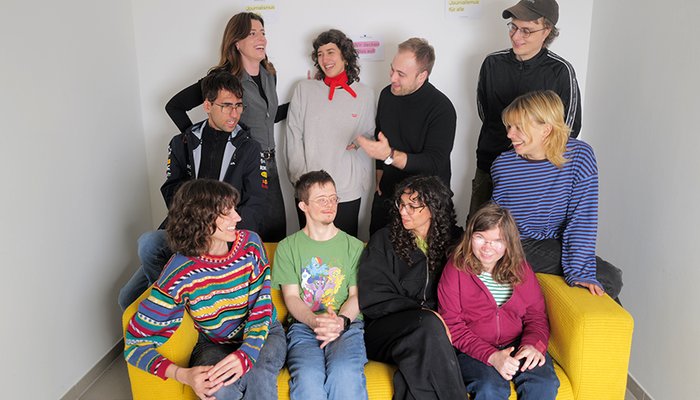10 Männer und Frauen mit und ohne Behinderung sprechen miteinander. 5 Personen sitzen auf einer gelben Sofa, die anderen stehen., 
