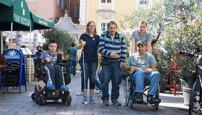 5 Personen vor schönen alten Häusern. Darunter 2 Rollstuhlfahrer:innen und eine blinde junge Frau.
