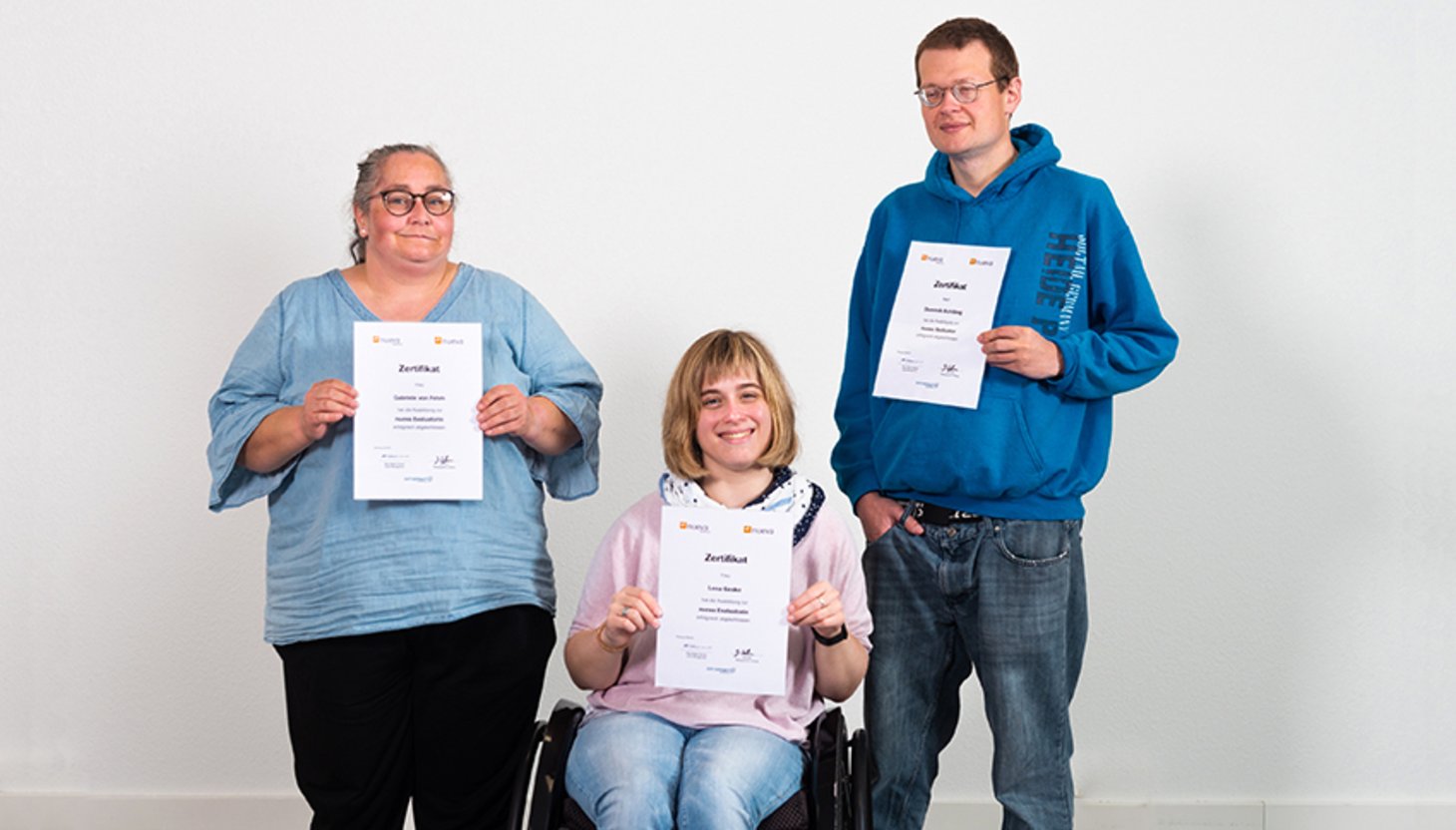 Eine Auszubildende mit Lernschwierigkeiten, eine Auszubildende im Rollstuhl und ein Auszubildender mit Sehbehinderung halten stolz ihre Zeugnisse in der Hand.