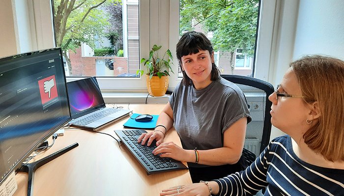 2 Frauen am Schreibtisch mit Bildschirm. Beide beraten sich, eine tippt auf der Tastatur.