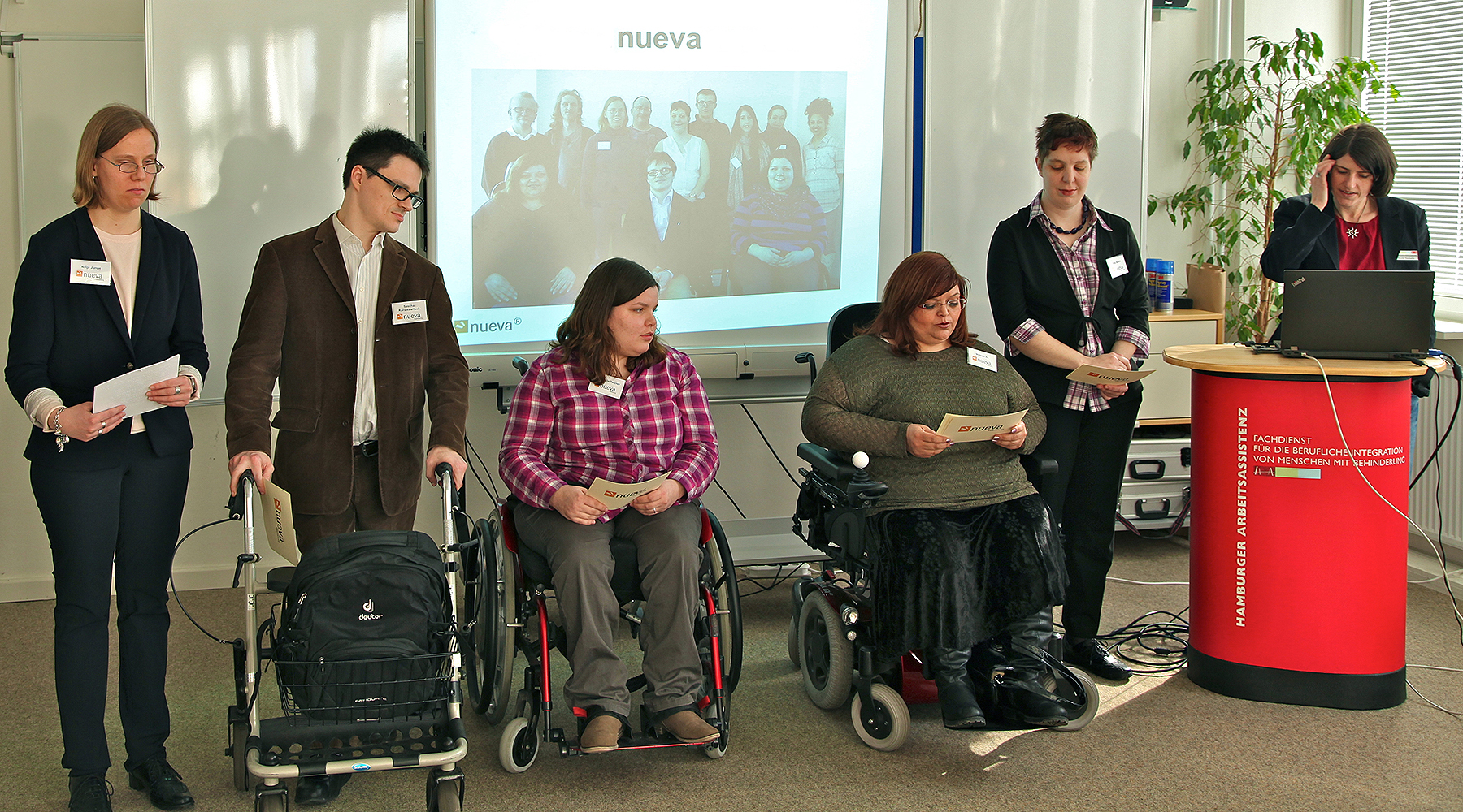 5 Team-Mitglieder von GUT GEFRAGT präsentieren nueva auf einer Veranstaltung. Die Gruppe besteht aus einem Evaluator und 3 Evaluatorinnen mit Behinderung und einer Mitarbeiterin am Pult.