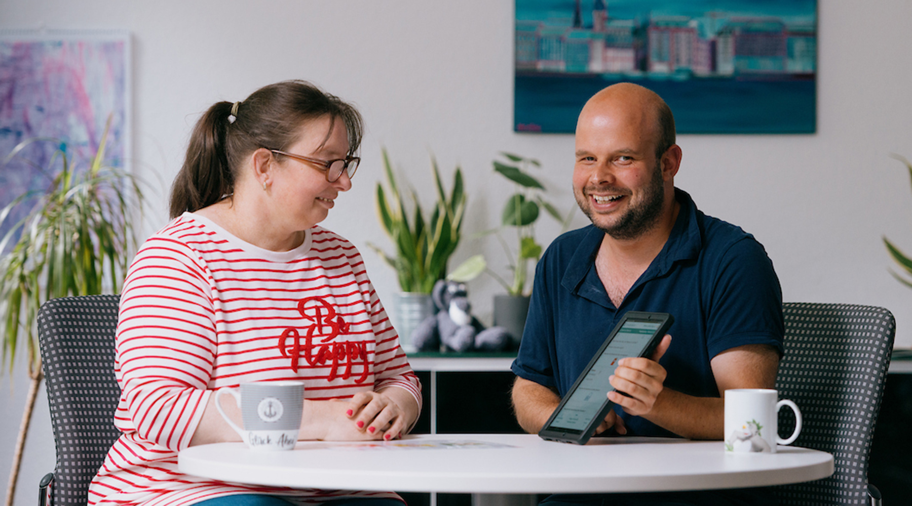 Eine Frau und ein Mann an einem Tisch, er hat ein Tablet in der Hand und lacht in die Kamera