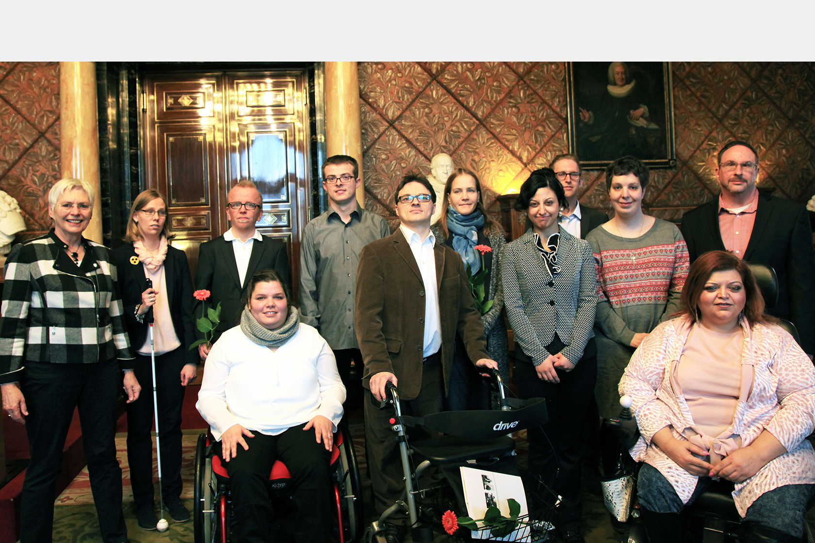 11 Auszubildende von GUT GEFRAGT bei der Übergabe ihrer Zertifikate im Hamburger Rathaus. Die Gruppe steht in einem prächtigen Saal mit Wandvertäfelung. Dazu gehören unter anderem zwei Rollstuhlfahrerinnen, ein Auszubildender mit Rollator und eine blinde Auszubildende.